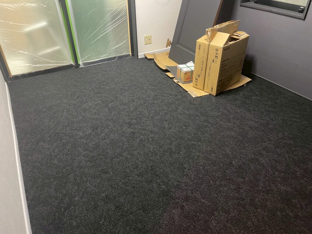 タイルカーペットを部屋全体に敷設している様子。濃いグレーのカーペットで落ち着いた印象の室内に仕上げている途中。